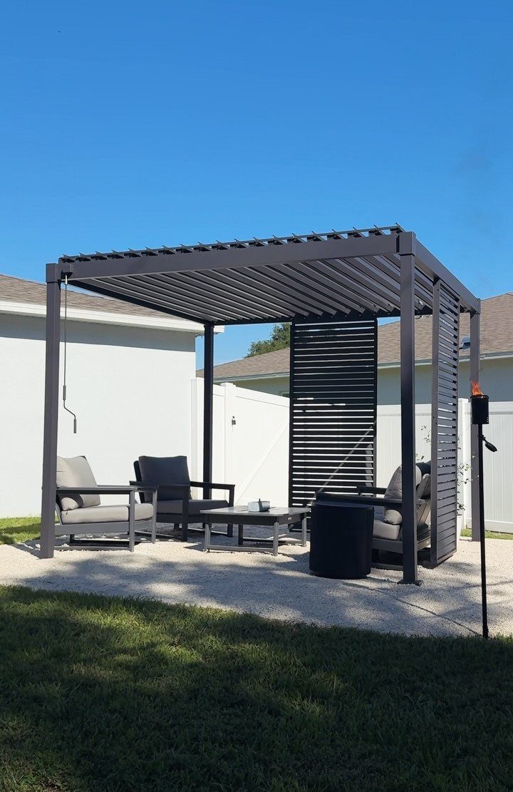 13 Backyard Entertaining Area Ideas for Every Season 1 Grey cushioned lounge chairs and a coffee table sit under a dark metal pergola.
