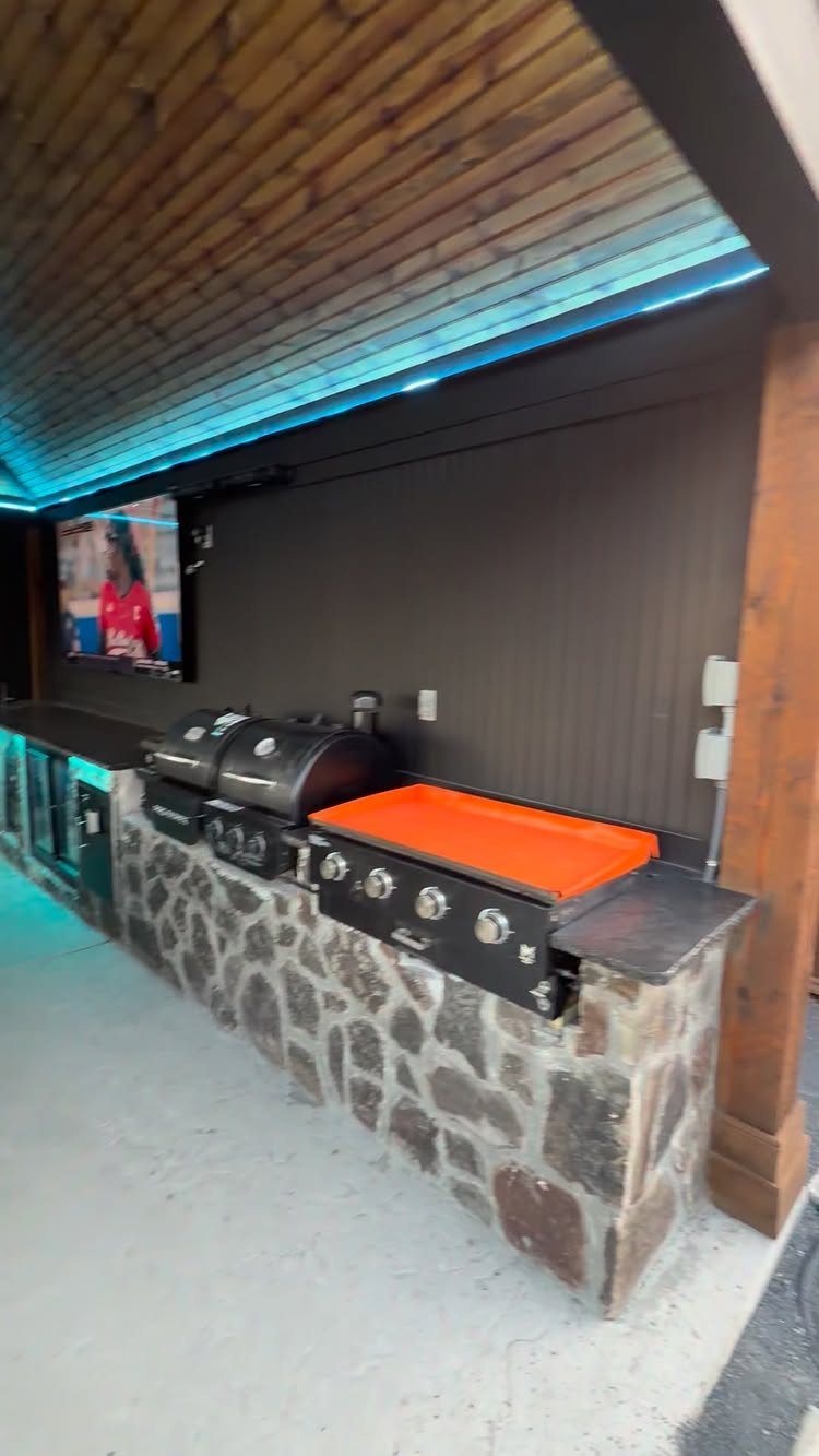 13 Backyard Entertaining Area Ideas for Every Season 4 Outdoor kitchen with stone base, black countertops, built-in grill, and television under blue LED lighting.