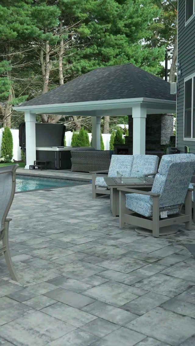 13 Backyard Entertaining Area Ideas for Every Season 12 Stone patio with blue cushioned chairs overlooking a swimming pool and hot tub under a pavilion.
