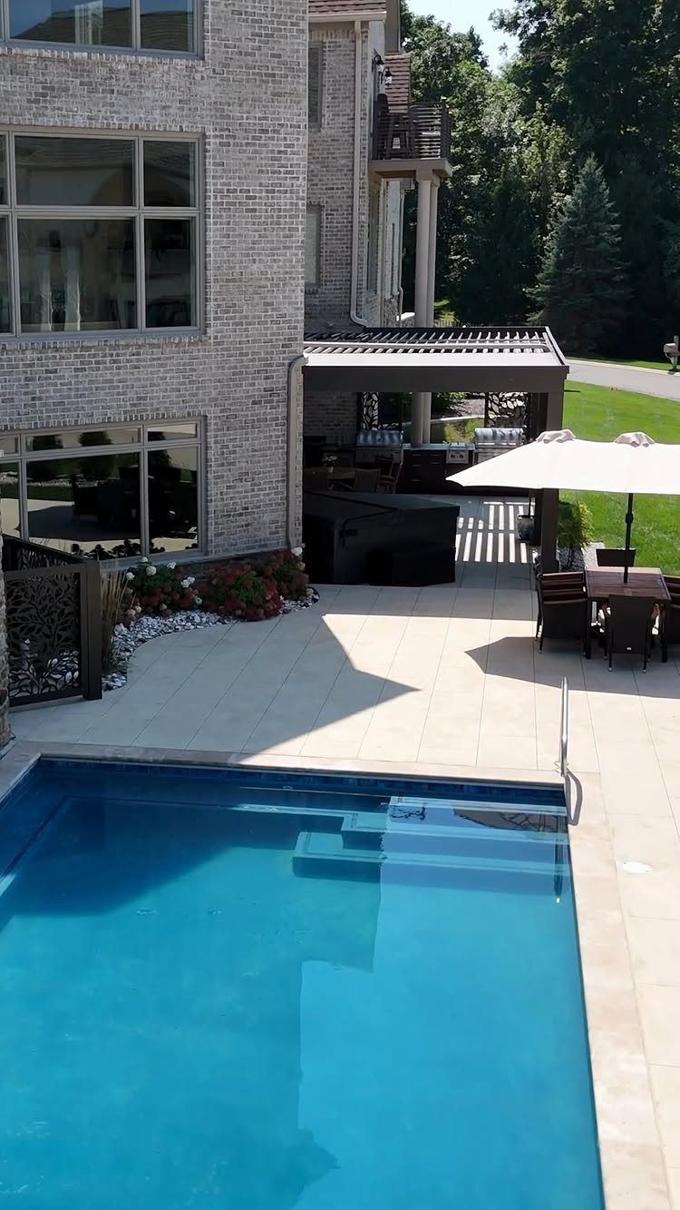 13 Backyard Entertaining Area Ideas for Every Season 3 A stone patio with a swimming pool, dining area, and a dark pergola shade structure.