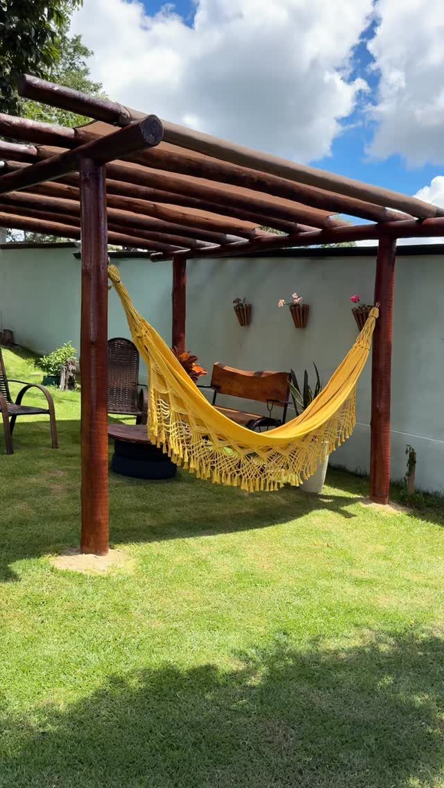 A yellow fringed hammock hanging under a wooden pergola in a sunny green backyard.