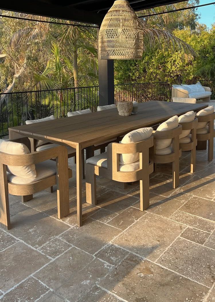 Wooden dining table and cushioned armchairs set on a stone patio surrounded by tropical greenery.