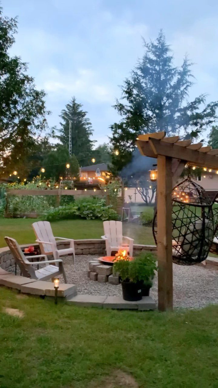 A stone fire pit with a burning fire surrounded by white chairs in a backyard.