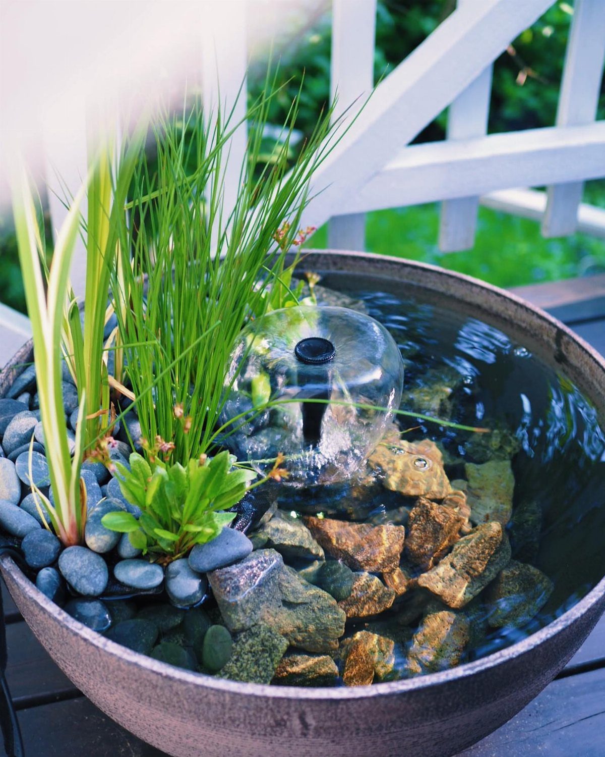 A small stone bowl fountain with green plants, pebbles, and a bubbling water jet.