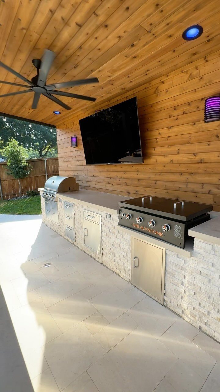 A modern outdoor kitchen with a stone island, stainless steel grill, and light tiled flooring under a wooden ceiling.