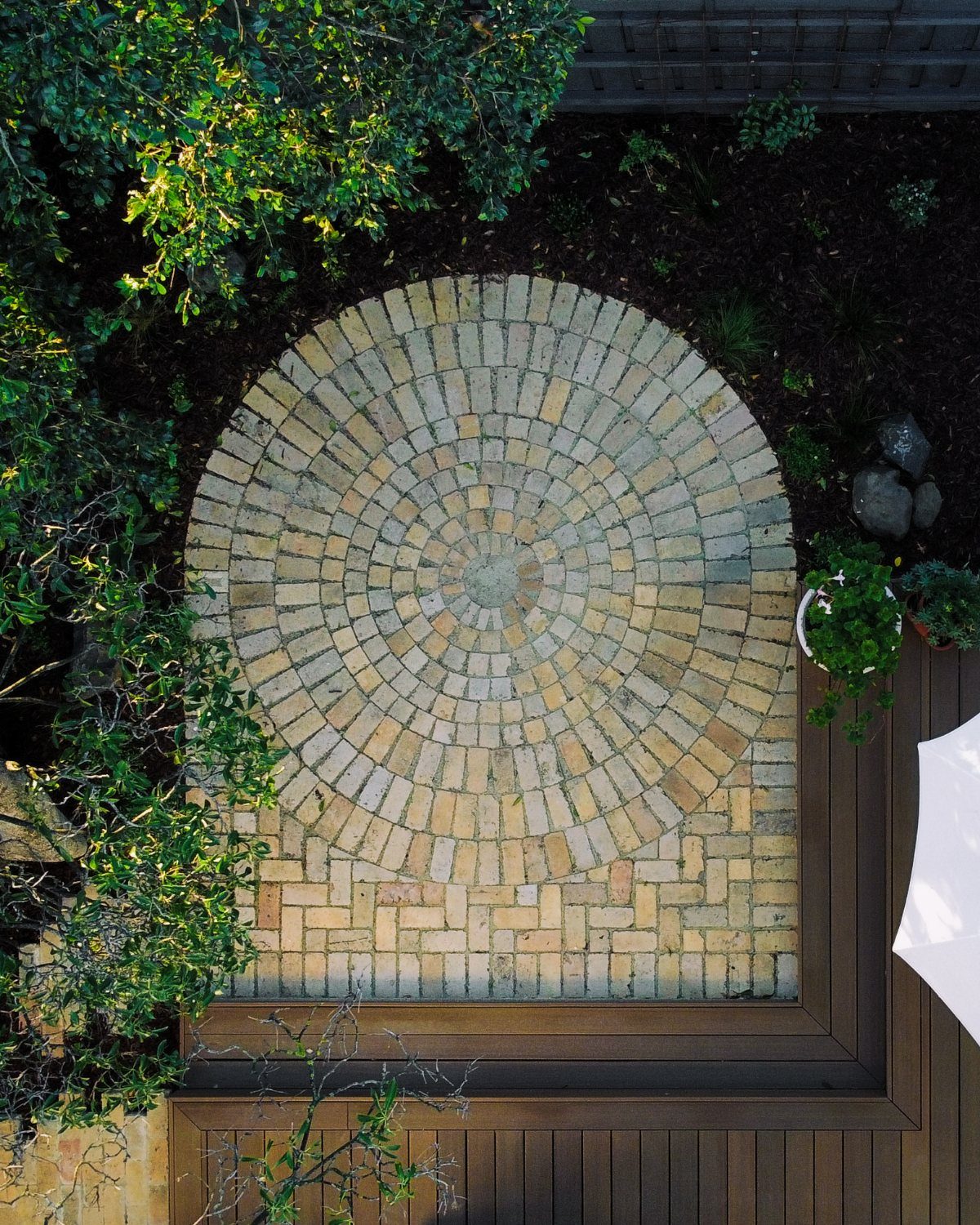 Circular brick patio with a fan-like radiating pattern surrounded by green plants and dark soil.