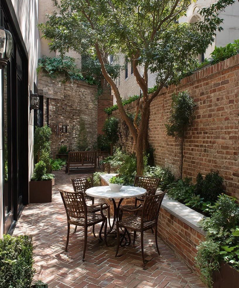 Red brick patio in a narrow courtyard with a small tree and outdoor dining set.