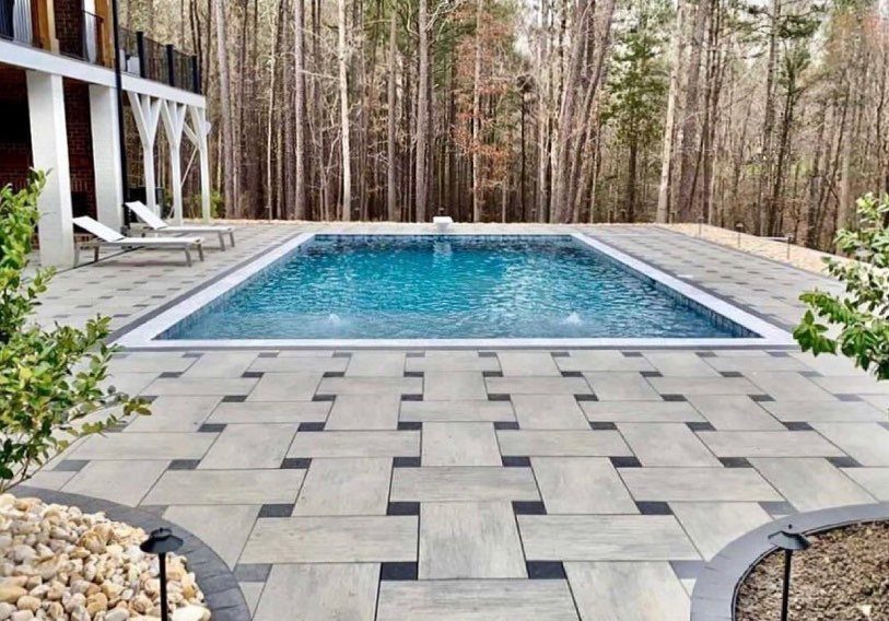 A geometric grey stone patio surrounds a rectangular blue swimming pool near a forest.