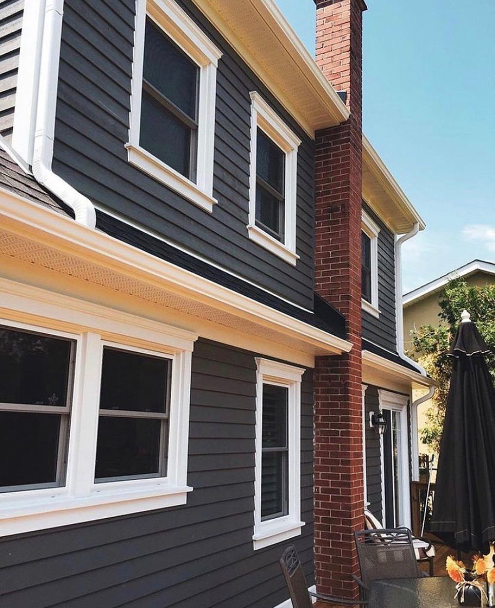 9 Cape Cod House Exterior Ideas for Curb Appeal 1 Dark grey house with white window trim and a red brick chimney against a blue sky.