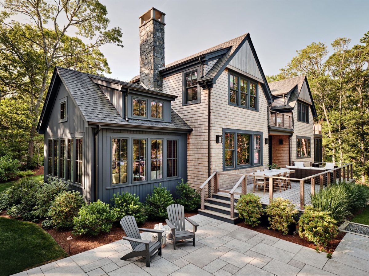 9 Cape Cod House Exterior Ideas for Curb Appeal 3 A large Cape Cod house with grey shingle shake siding and dark trim surrounded by trees.