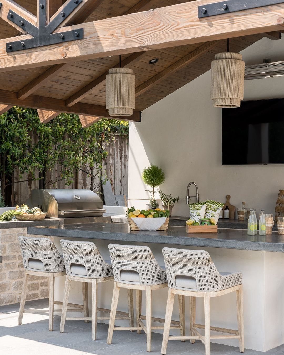 20 Covered Patio Ideas for Shade, Style and Year-Round Use 8 Outdoor kitchen with stone countertop, woven bar stools, and a wooden timber frame structure.