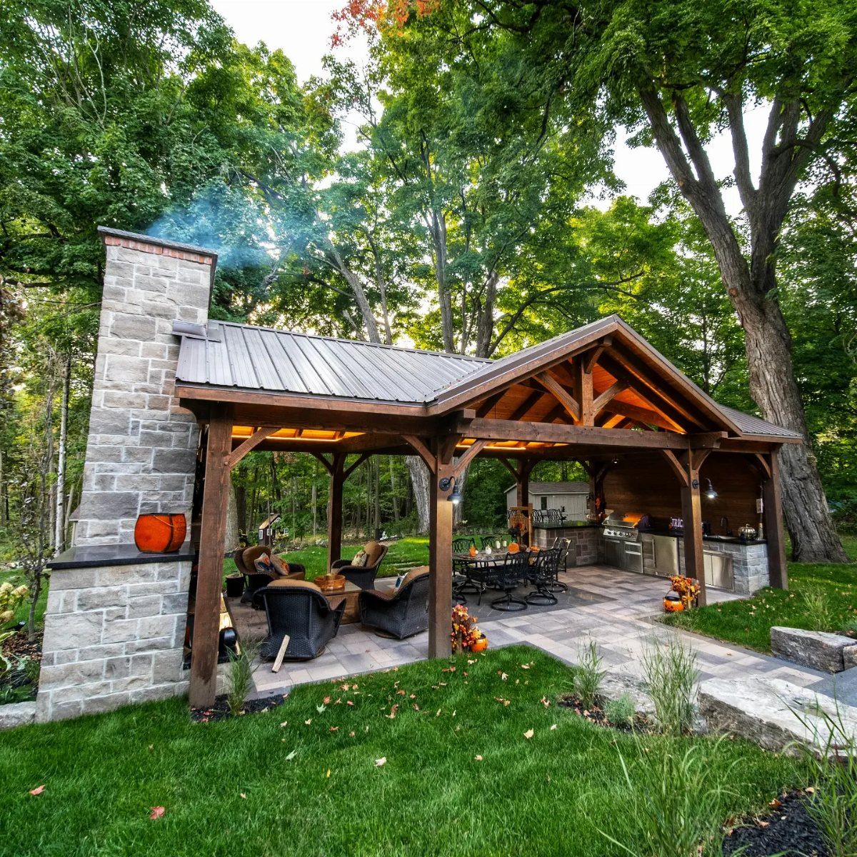 20 Covered Patio Ideas for Shade, Style and Year-Round Use 19 Timber frame pavilion with a stone fireplace, outdoor kitchen, and dining area in a wooded setting.