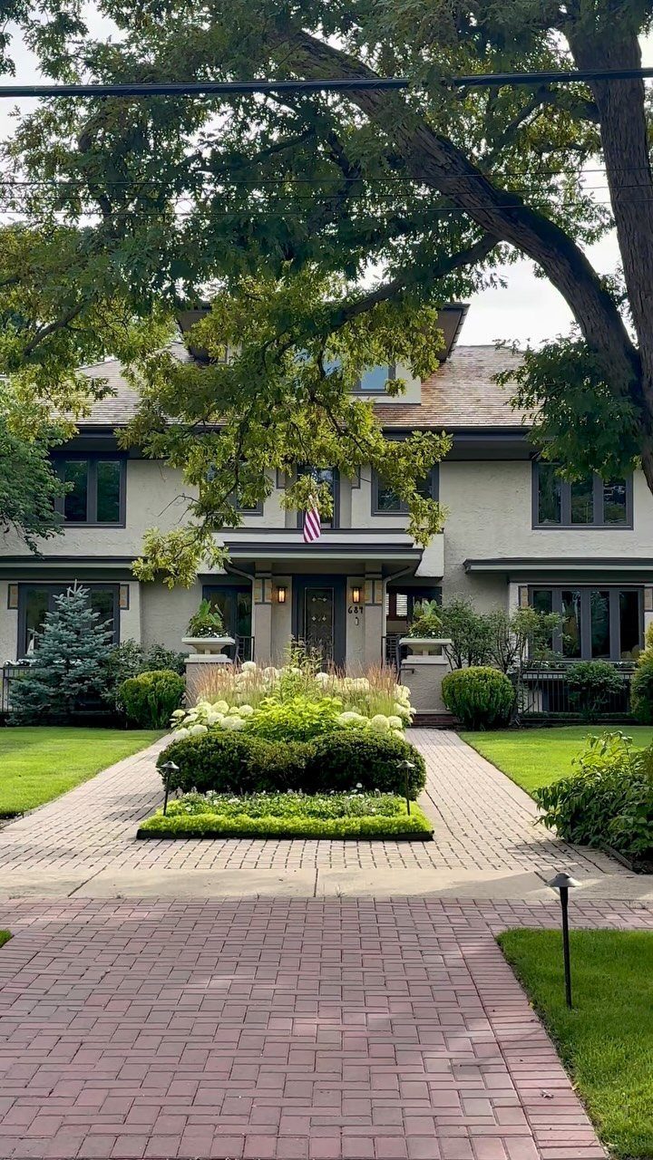 10 Curb Appeal Ideas for a Sharper Front Facade in 2026 8 A large grey house with dark windows, manicured gardens, and a brick walkway leading to the entrance.