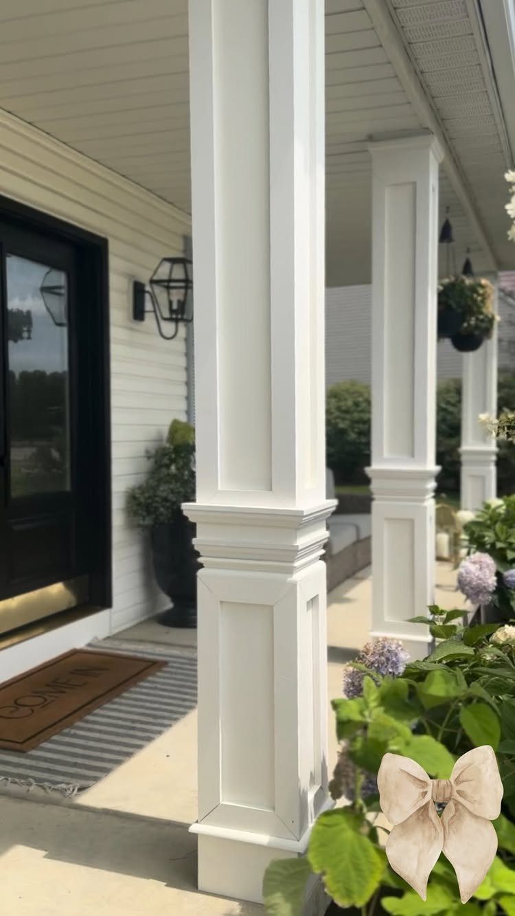 10 Curb Appeal Ideas for a Sharper Front Facade in 2026 6 White decorative paneled columns standing on a porch next to green plants and a black door.