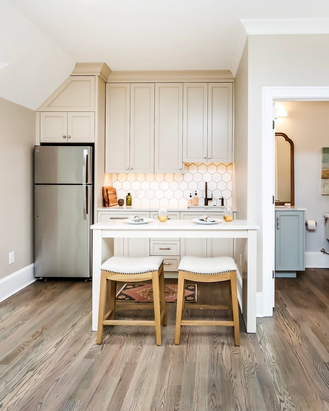Garage to Family Room Conversion: 8 Ideas for 2026 6 Small kitchenette with beige cabinets, white hexagonal backsplash, stainless steel refrigerator, and two wooden stools.
