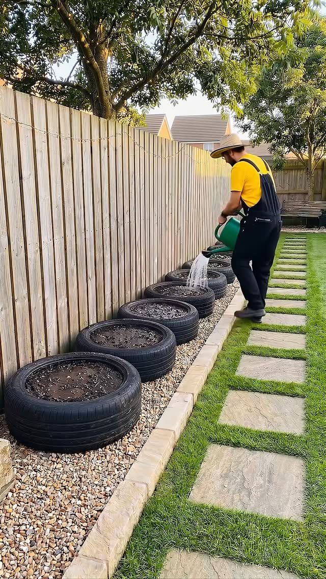 Man watering a row of black tires used as planters along a wooden fence.