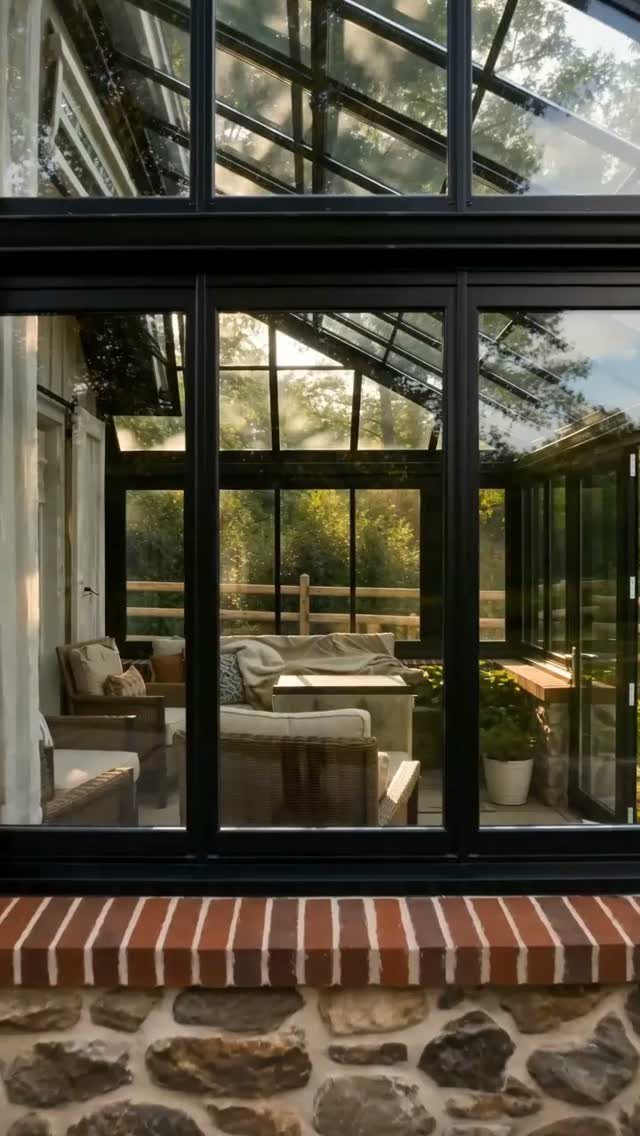 8 Houses With Greenhouses That Skip the Ordinary 3 A glass sunroom with black frames and comfortable seating overlooking a lush green forest.