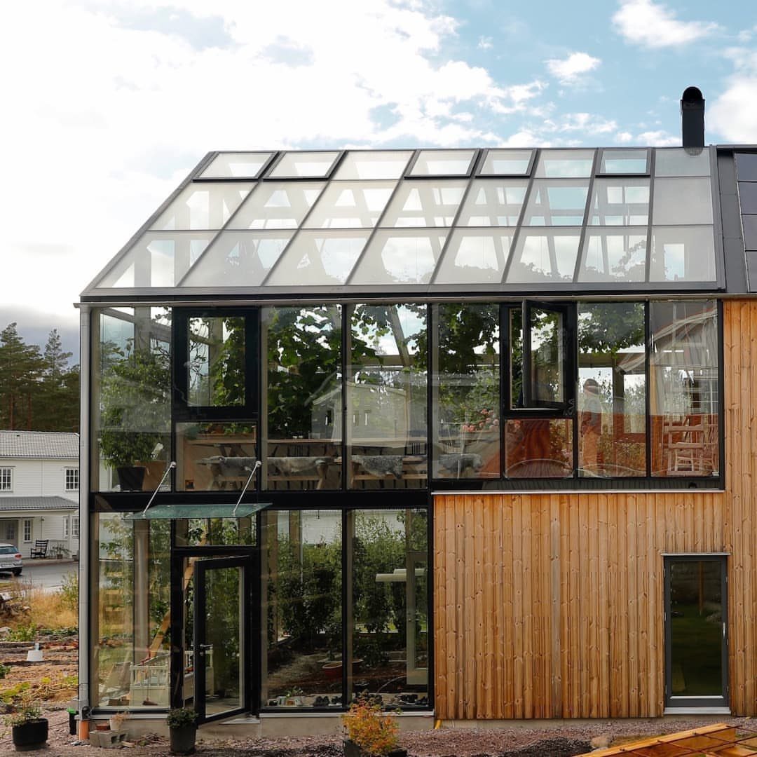 8 Houses With Greenhouses That Skip the Ordinary 6 A modern house with a large integrated glass greenhouse structure and wooden siding under a cloudy sky.