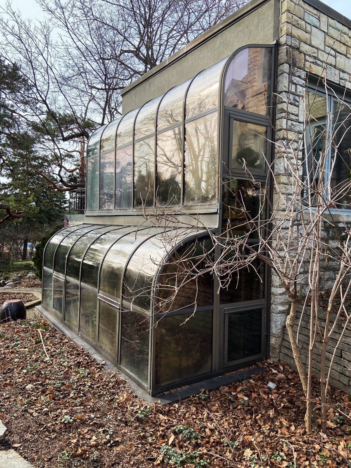 8 Houses With Greenhouses That Skip the Ordinary 5 A two-tiered glass greenhouse structure attached to a stone house surrounded by bare trees.