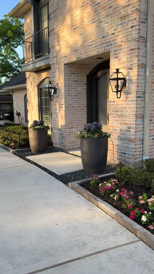 A brick house entrance with large planters and a small flower bed along a concrete walkway.
