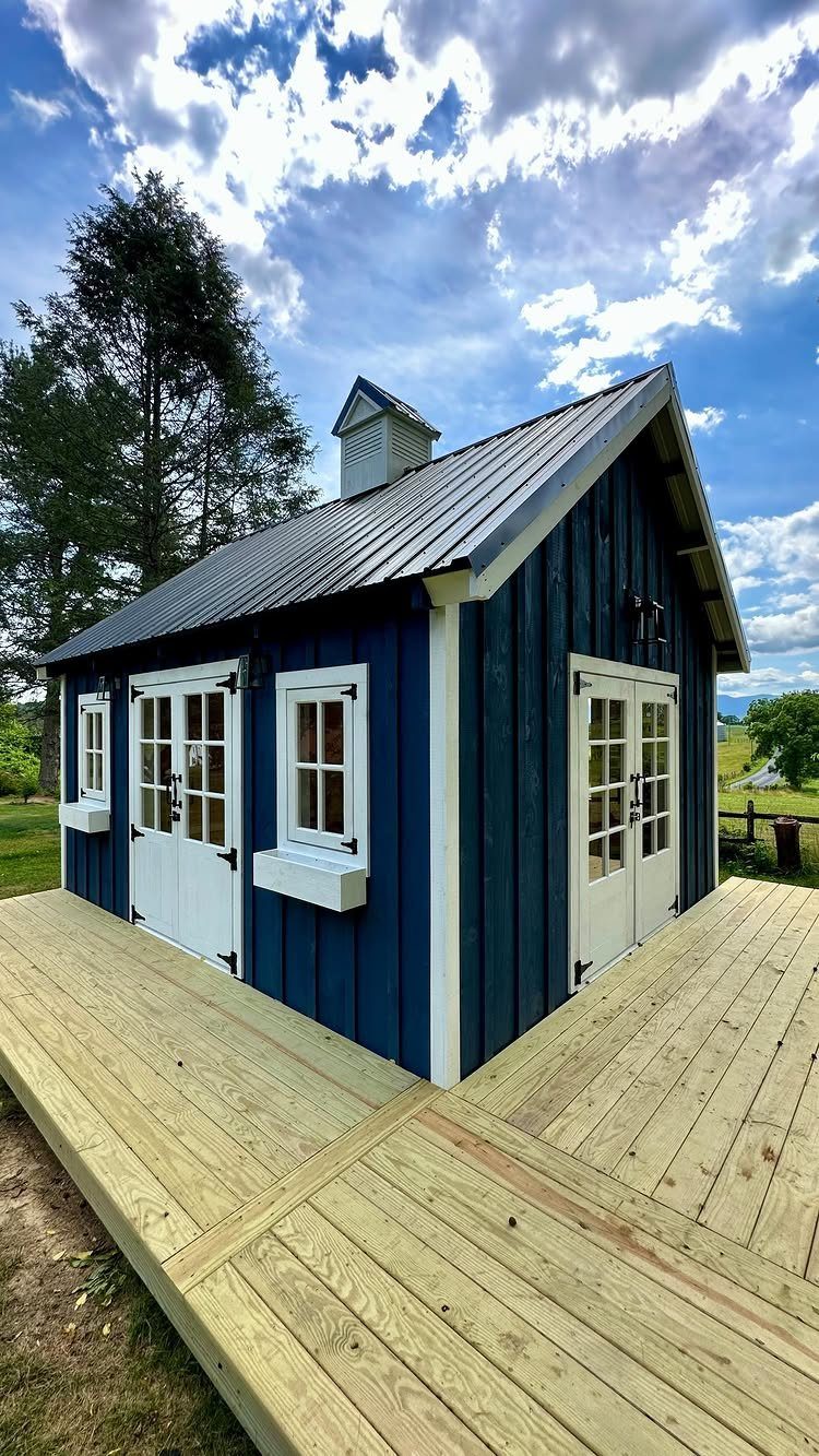 9 Modern Farmhouse Shed Ideas for Your Backyard 9 A blue modern farmhouse style shed with white trim and a metal roof on a wooden deck.