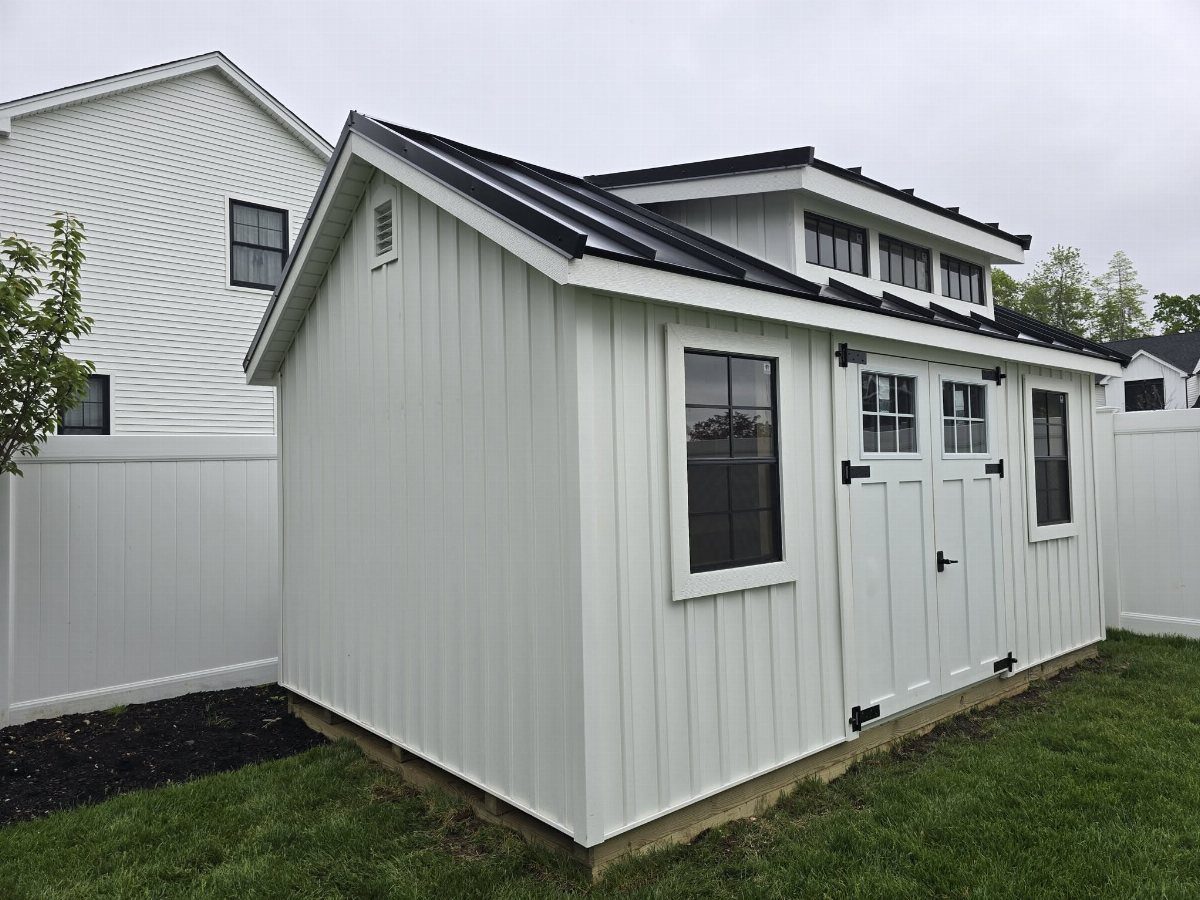 9 Modern Farmhouse Shed Ideas for Your Backyard 1 White board and batten shed with a black metal roof and dark window trim on a lawn.