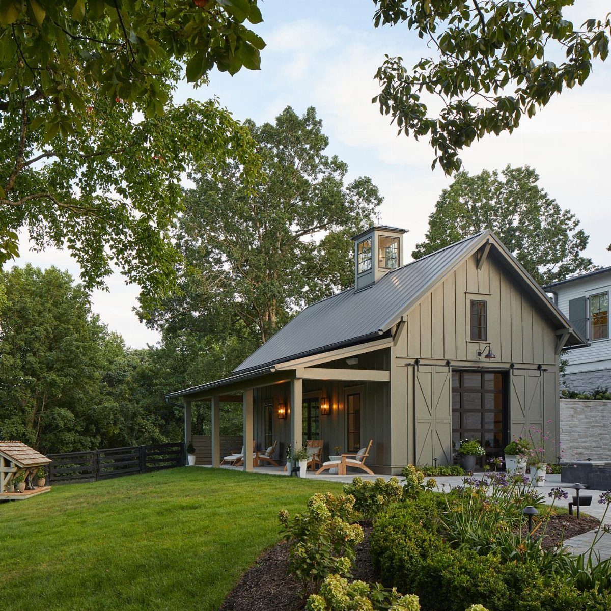 9 Modern Farmhouse Shed Ideas for Your Backyard 6 Olive green farmhouse garden shed with a large covered porch and dark metal roof in a garden.