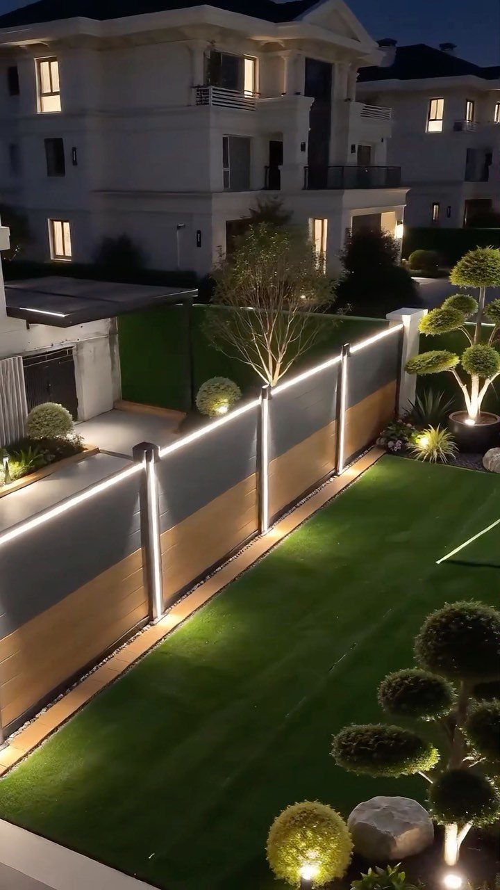22 Outdoor Backyard Ideas for Dining, Gardens, and Gathering Spaces 19 Modern backyard fence with integrated LED light strips bordering a green lawn at night.