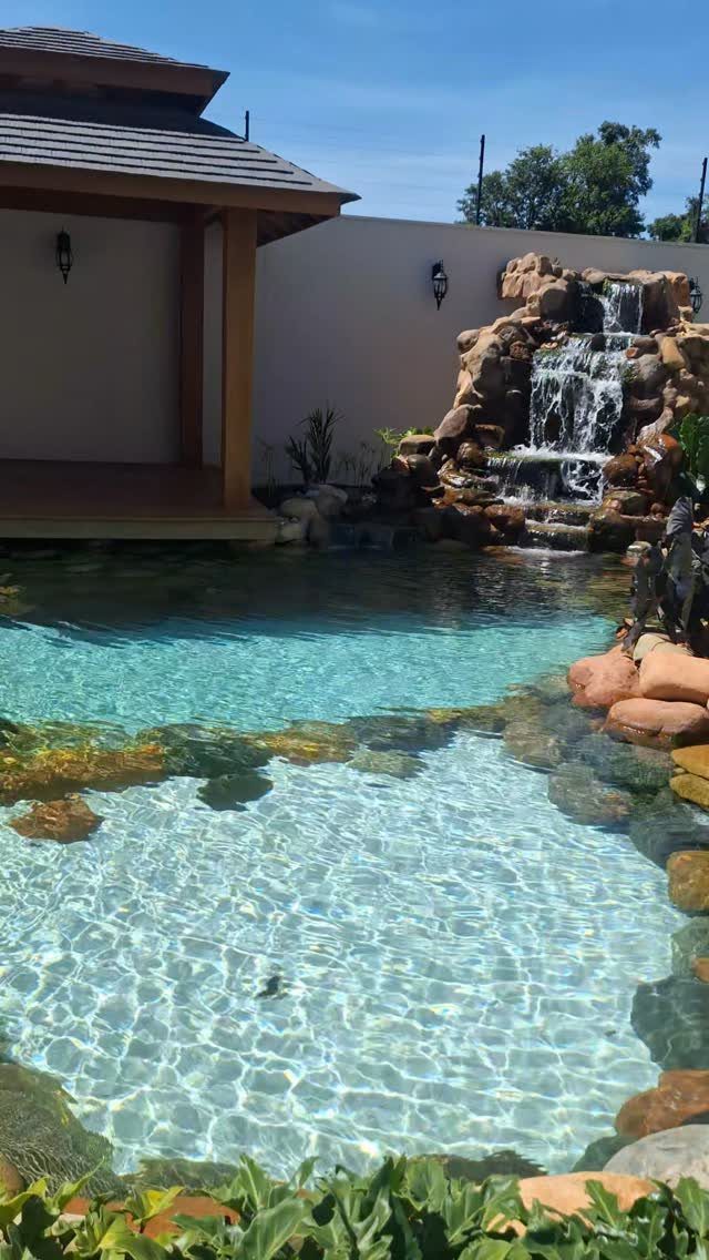 22 Outdoor Backyard Ideas for Dining, Gardens, and Gathering Spaces 16 A clear blue swimming pool with a natural rock waterfall and lush green plants.