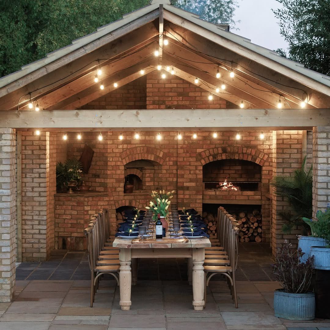 22 Outdoor Backyard Ideas for Dining, Gardens, and Gathering Spaces 6 A long wooden dining table set for a meal under a lit brick pavilion.
