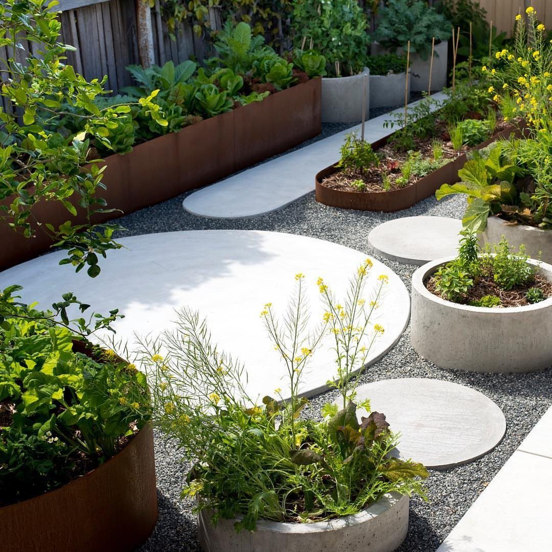 29 Outdoor Gardens Ideas for Patios, Yards, and Every Budget 3 Edible plants growing in rusted metal raised beds and concrete planters amidst grey gravel.
