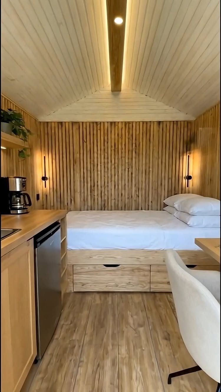 Small wooden interior with a bed, kitchenette, and light wood paneled walls and ceiling.