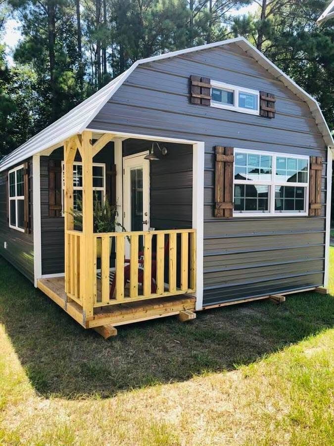 A grey tiny shed house with a wooden porch and white-trimmed windows in a grassy yard.