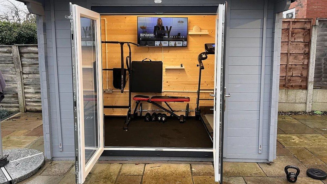 A grey garden shed converted into a home gym with black rubber mats on the floor.