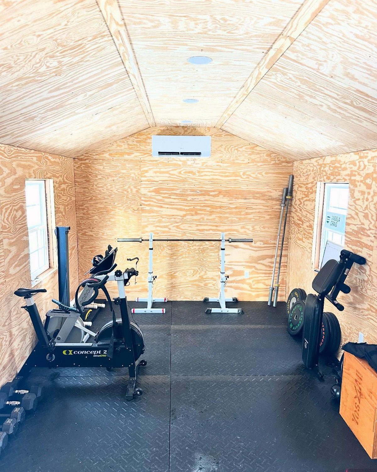 Gym equipment including a rowing machine and barbell inside a wooden shed with rubber mats.